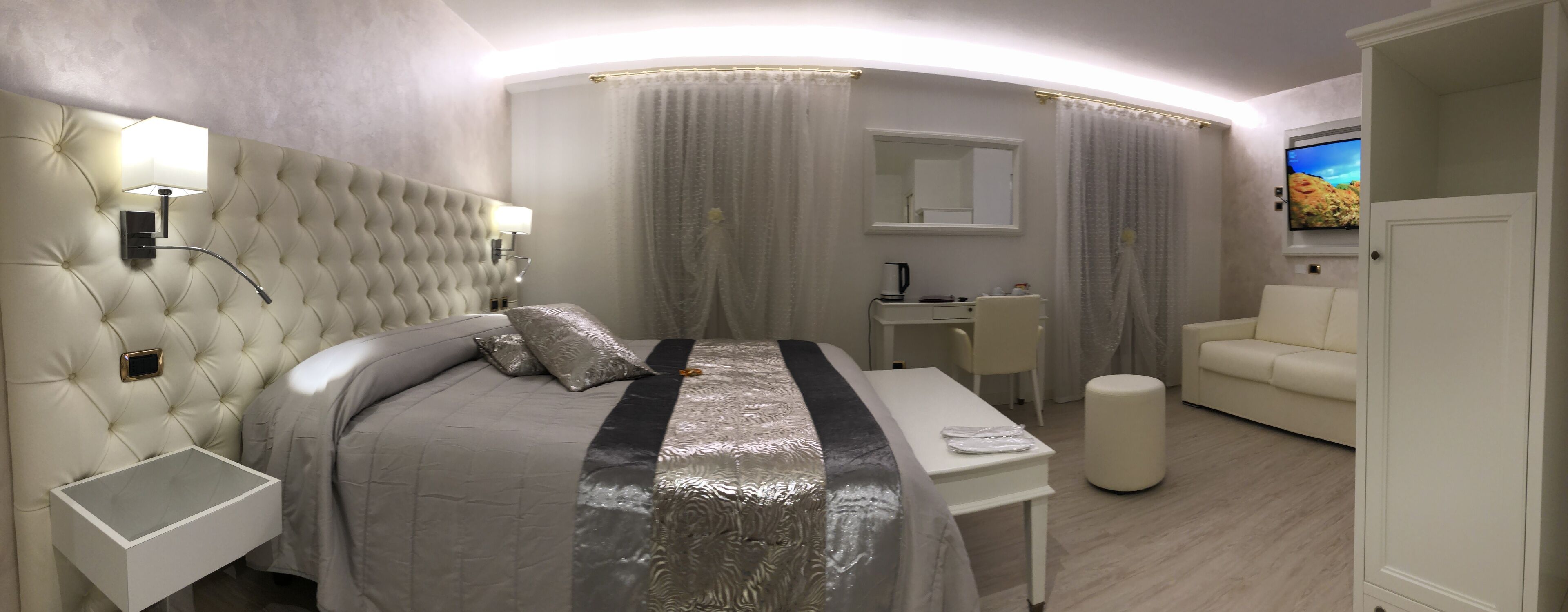 A modern hotel room with a tufted headboard, a bedside table, a bed with a metallic bedspread, a small desk with a chair, a white sofa, and a television on the wall.