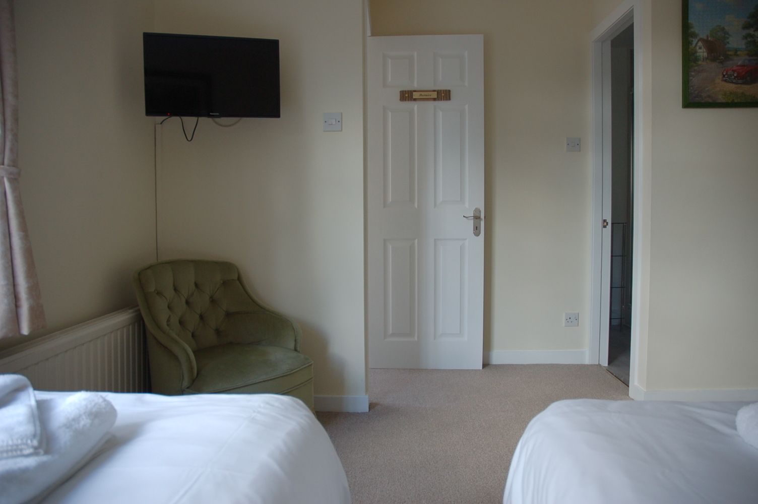 A hotel room with two beds, a television mounted on the wall, a door with a nameplate, and a painting on the wall.