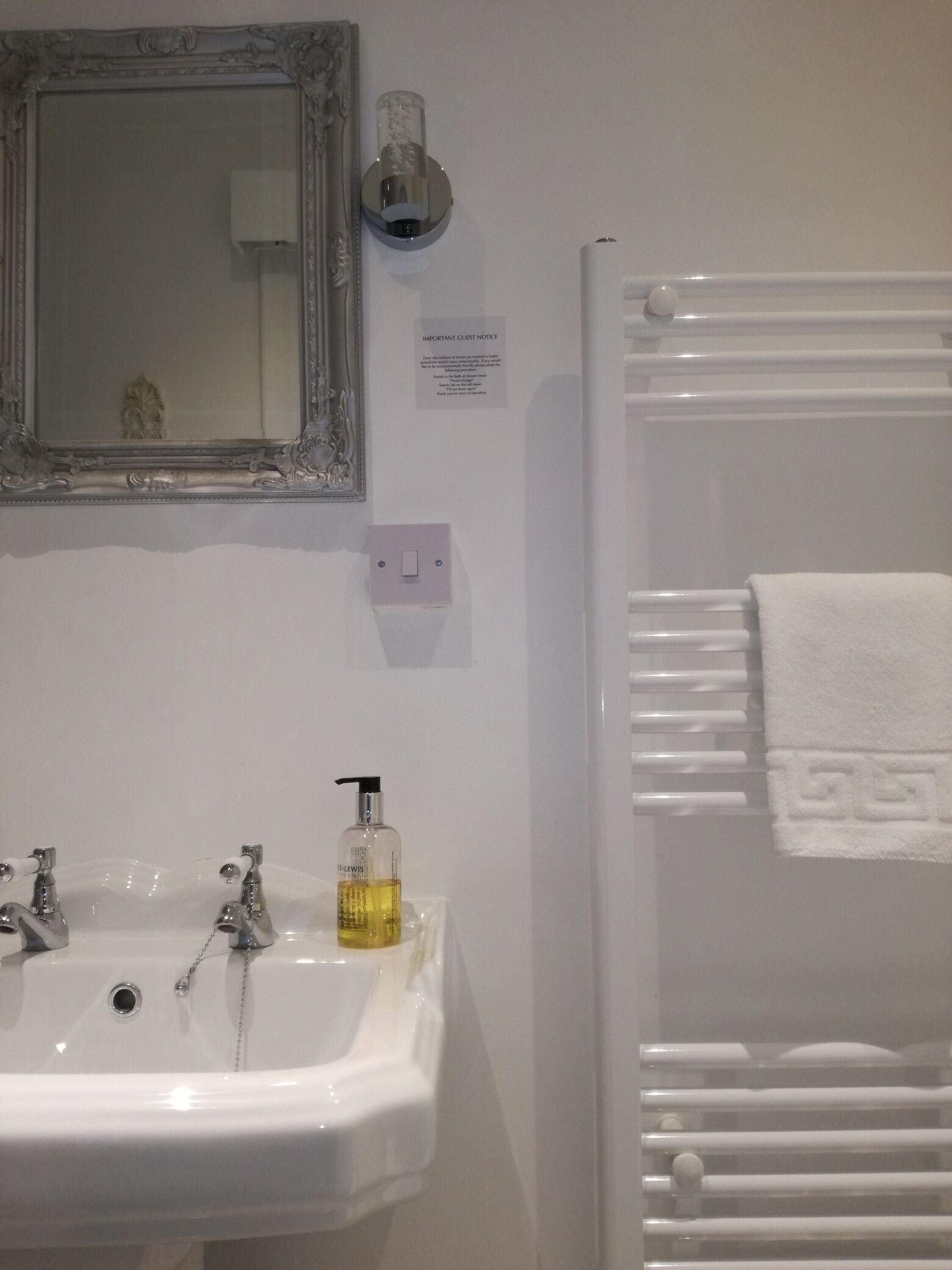 A bathroom with a white sink, two faucets, a mirror with an ornate frame, a wall-mounted light, and a towel rack with a white towel.