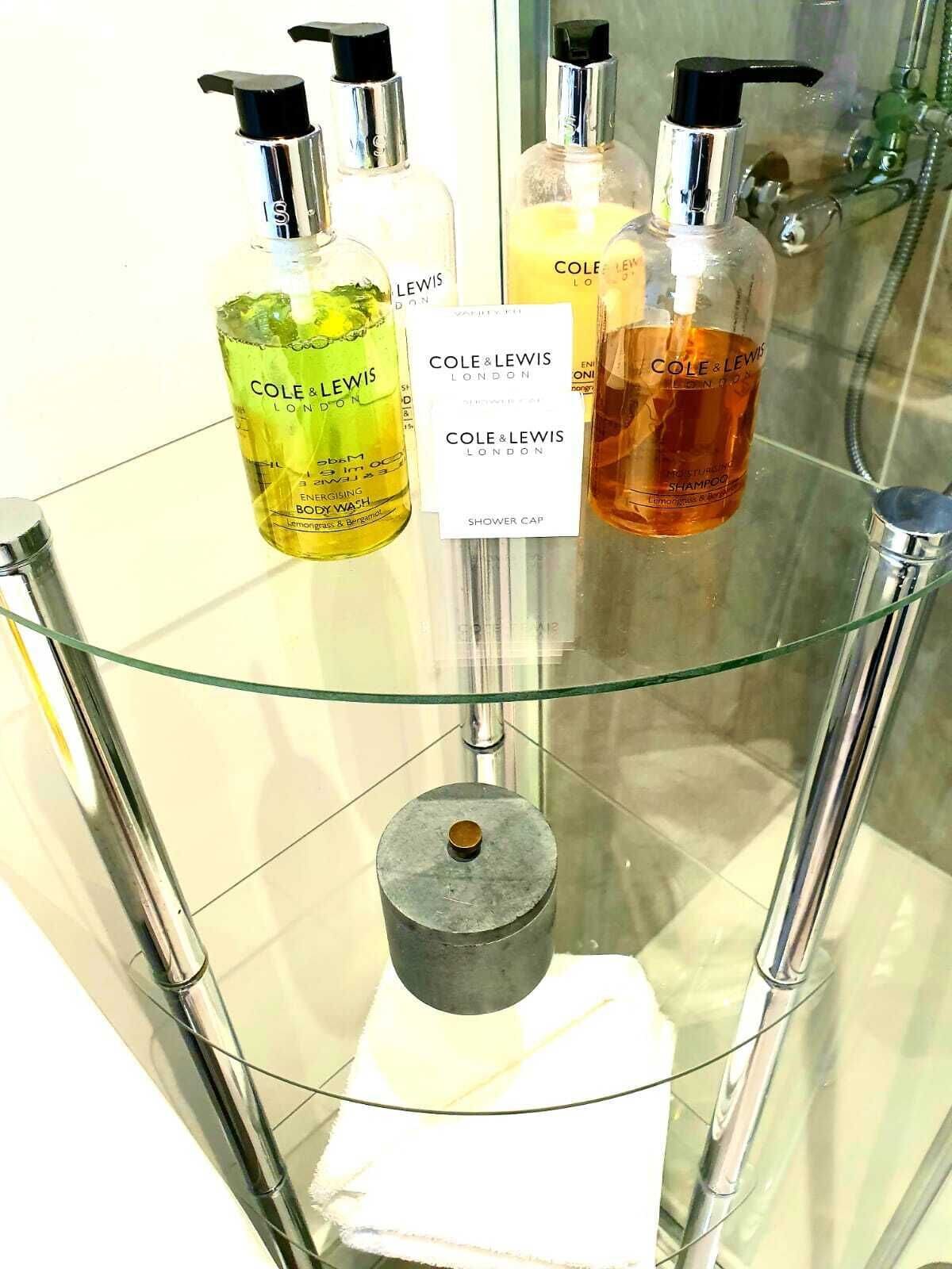 A glass shelf with bottles of Cole & Lewis London products, including a shower cap and shampoo.