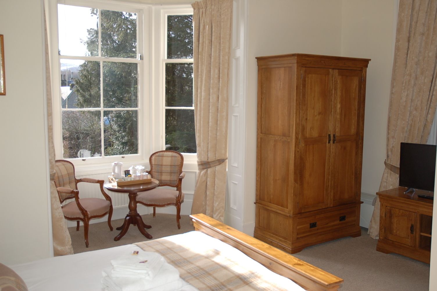 Une chambre à coucher avec un lit, une armoire en bois, un téléviseur posé sur un meuble en bois et une fenêtre avec des rideaux.