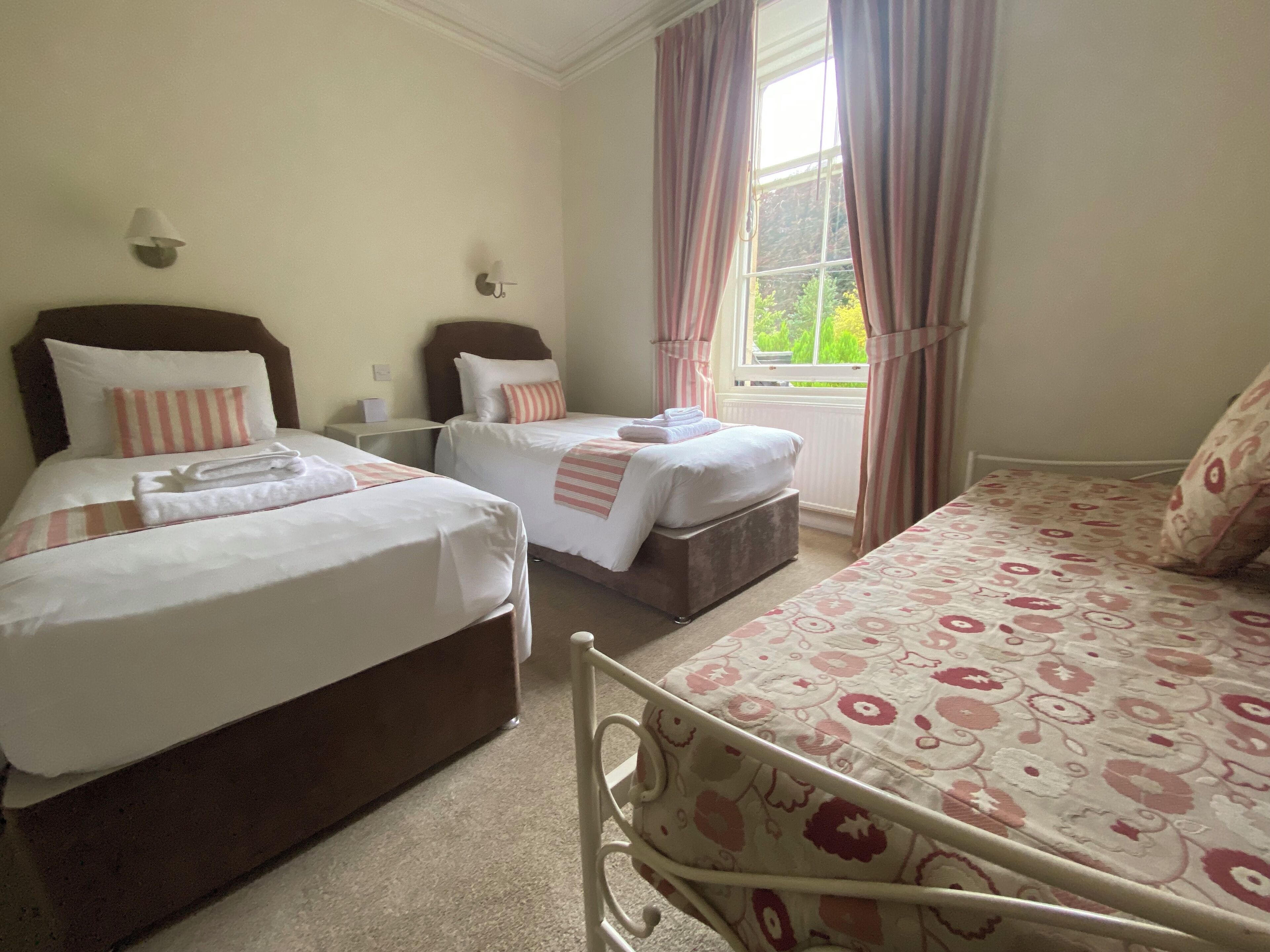 A hotel room with two single beds, a large window with curtains, a bedside table, and a wall-mounted lamp.
