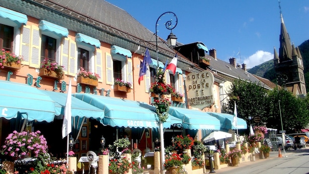 Une rangée de bâtiments aux couleurs vives, dotés d’auvents bleus, dont certains sont ornés de jardinières et de drapeaux.