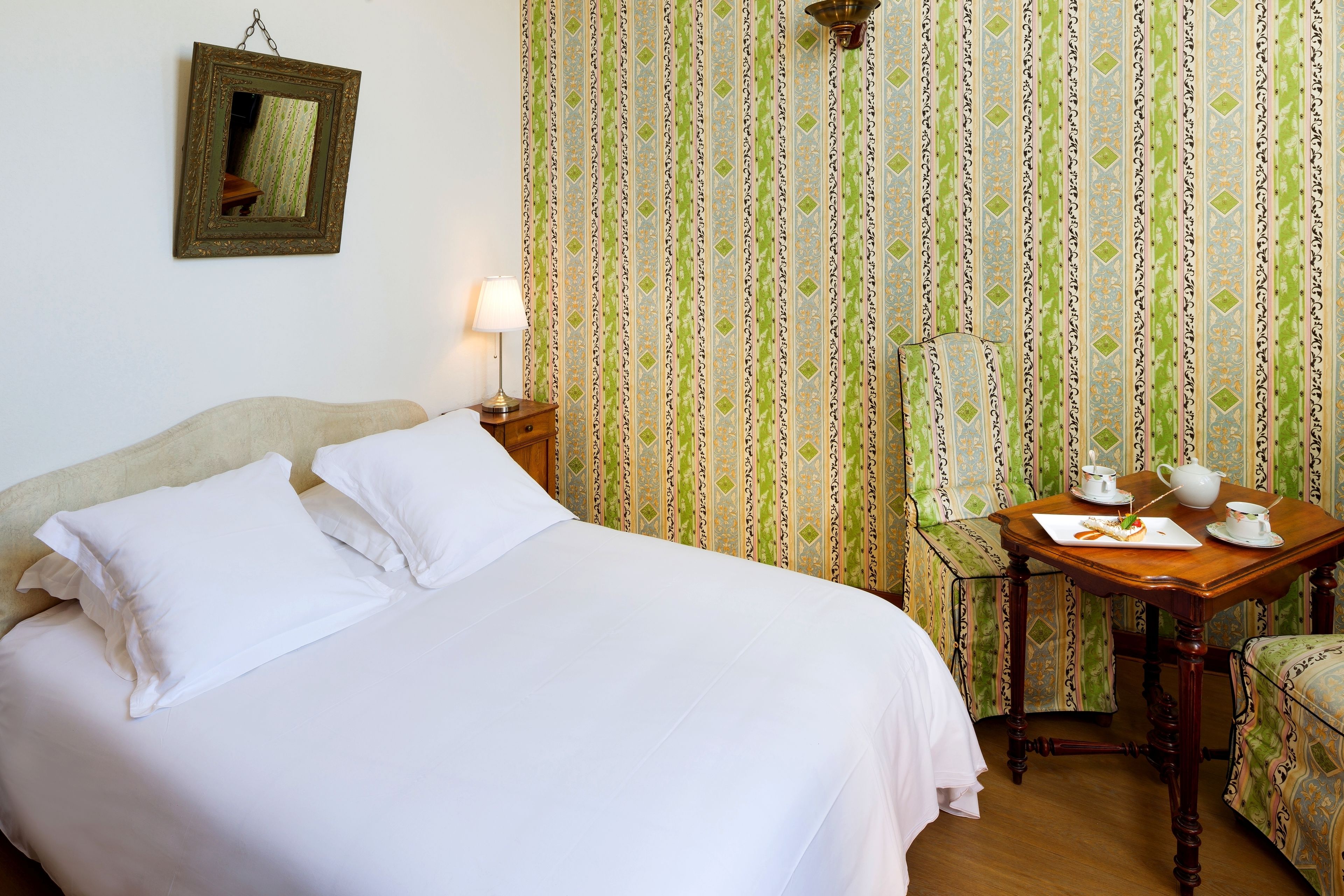 A neatly made bed with white linens, a bedside table with a lamp, a chair, and a small table with a teapot and cups.