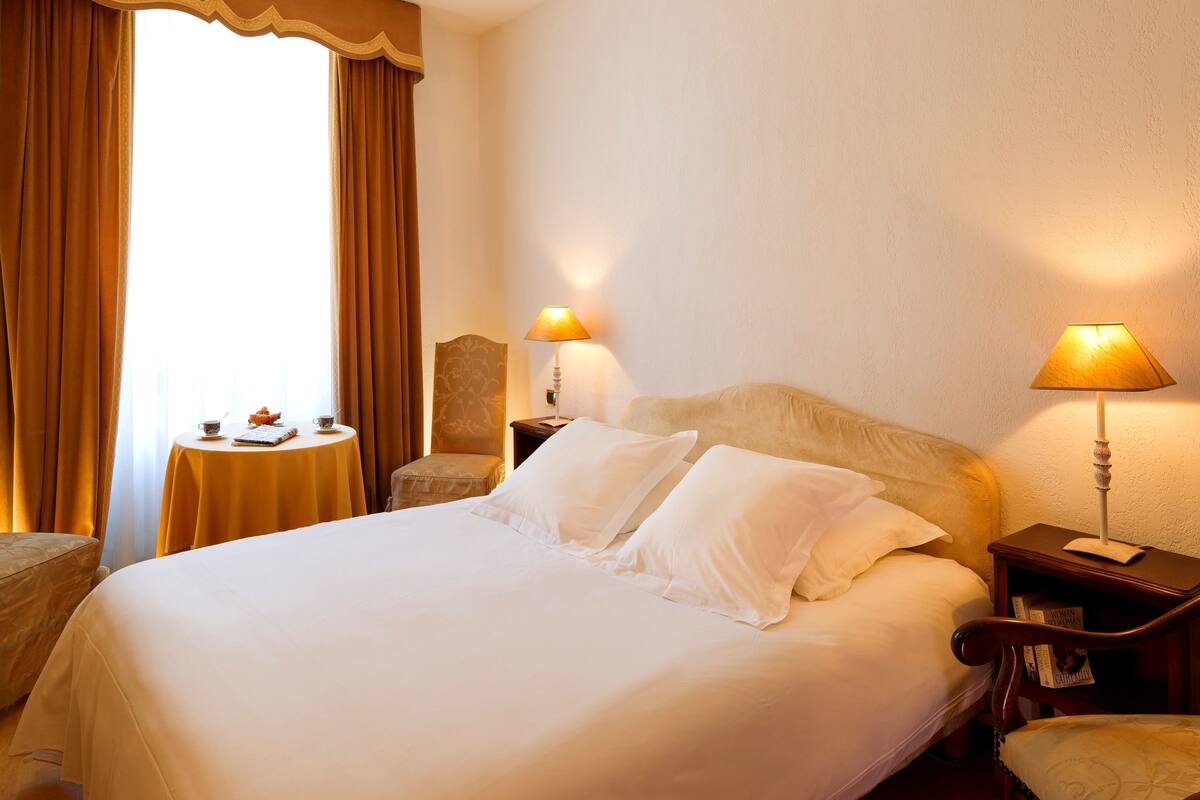 A hotel room with a large bed, two bedside lamps, a small round table with a chair, and a window with curtains.