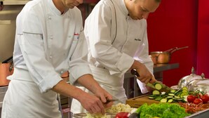 Deux chefs préparent des plats dans une cuisine dont le mur est rouge et où l’on peut voir des casseroles en cuivre.