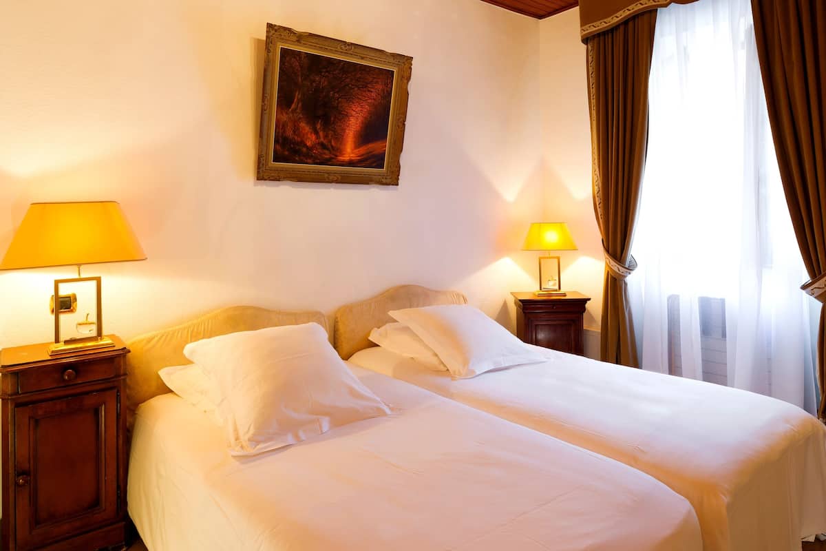 A hotel room with two beds, a nightstand with a lamp, a framed picture on the wall, and a window with curtains.