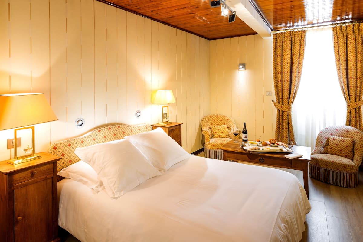 A hotel room with a bed, two bedside tables with lamps, a small table with a bottle and food, and two patterned armchairs.