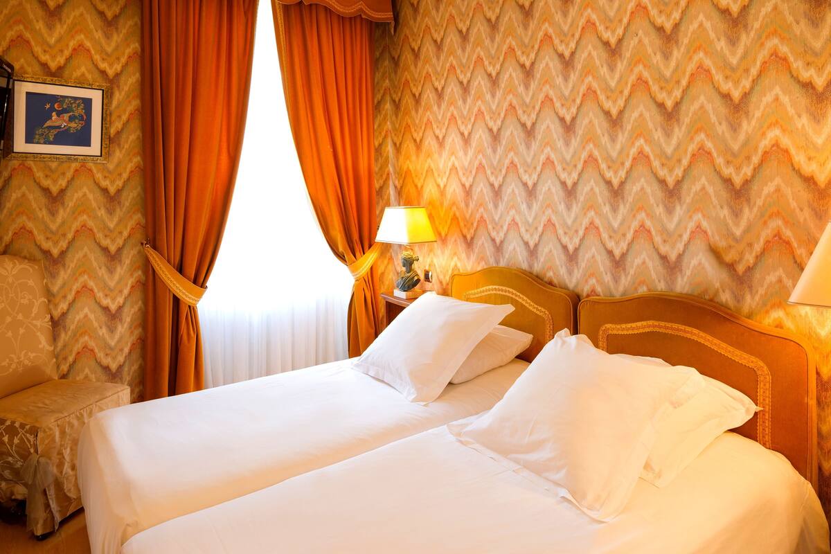 A hotel room with two beds, a nightstand with a lamp, and a window with orange curtains.