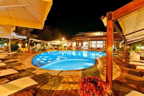 Un área de piscina con sillones de descanso y sombrillas, rodeada por una terraza de madera y un edificio con grandes ventanales.