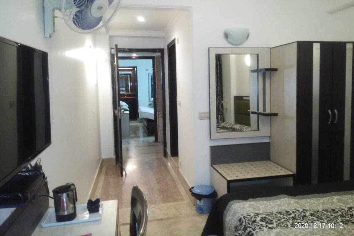 A hotel room with a flat-screen TV, a standing fan, a large mirror, a wardrobe, a small table with a kettle, and a bed.