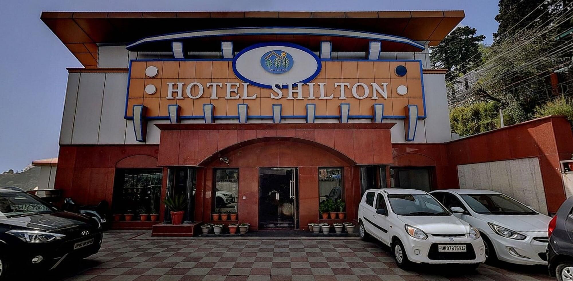 Entrada de hotel com uma placa indicando "HOTEL SHILTON" e um logotipo acima. Vários carros estão estacionados em frente.