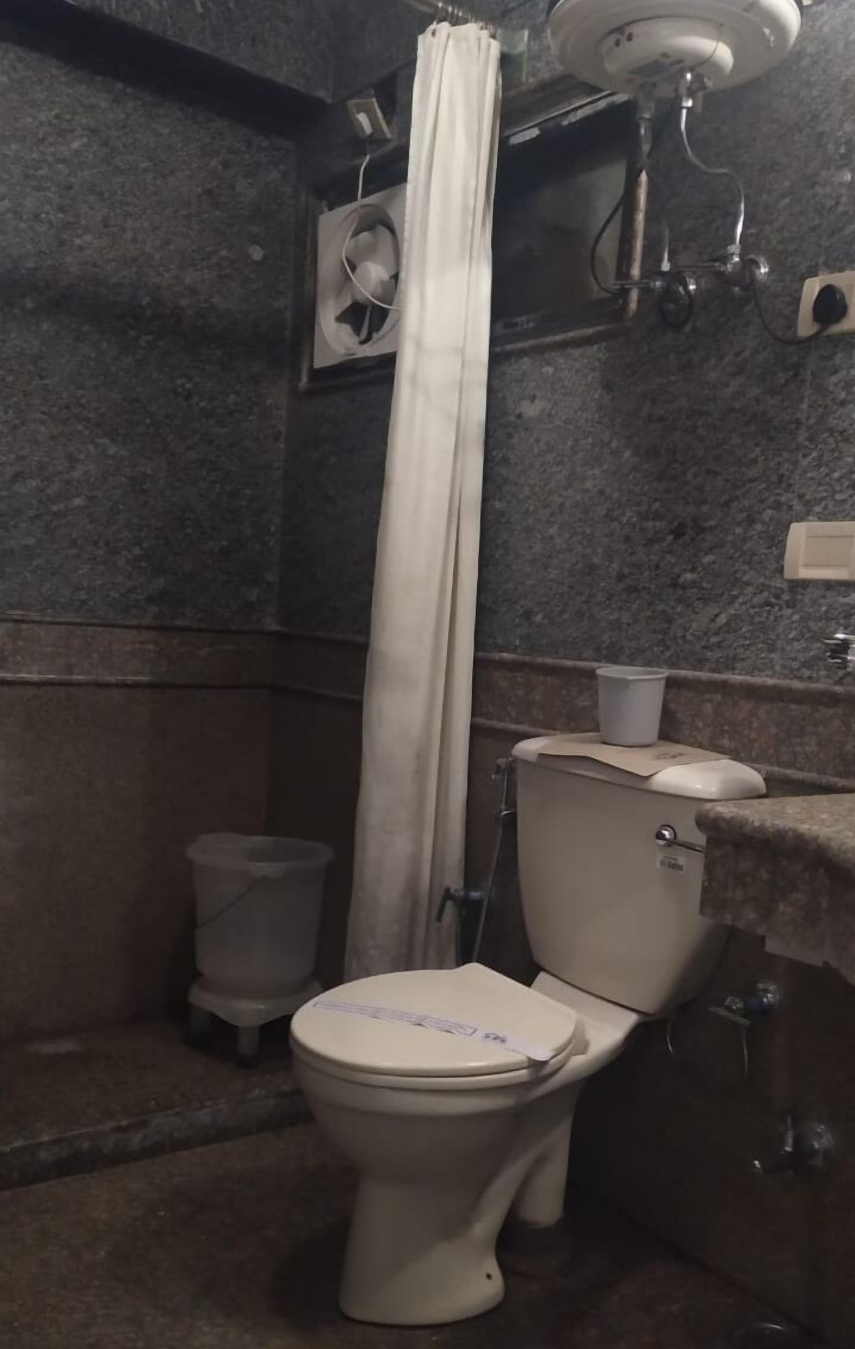 A bathroom with a toilet, a shower curtain, a wall-mounted fan, and a trash bin.