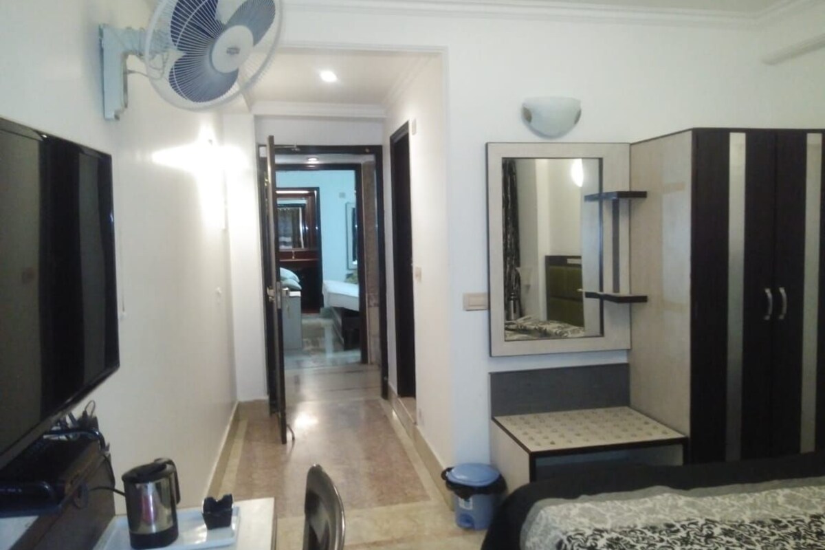 Une chambre d’hôtel équipée d’une télévision à écran plat, d’un ventilateur sur pied, d’un grand miroir, d’une armoire, d’une petite table avec une bouilloire et d’un lit.