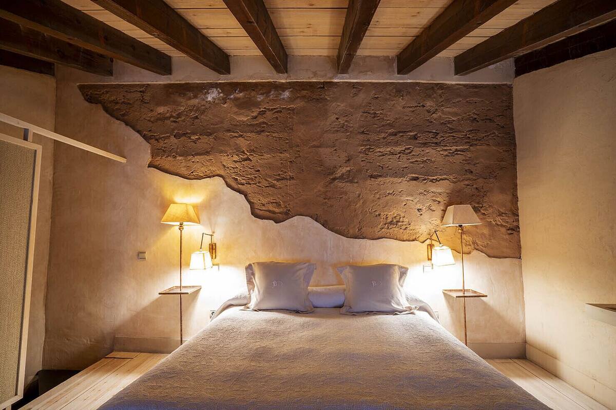 Een slaapkamer met een rustiek stenen plafond, een bed met wit beddengoed en twee bedlampen.