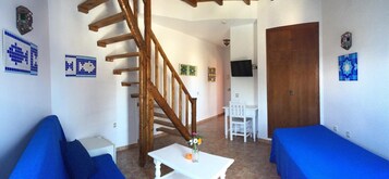 Una habitación con una escalera de madera, una mesa blanca con un jarrón, sofás azules, una silla blanca y una puerta de madera.