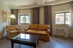 A room with a sofa, armchair, and a small table. There are windows with curtains, a lamp, and framed pictures on the wall.