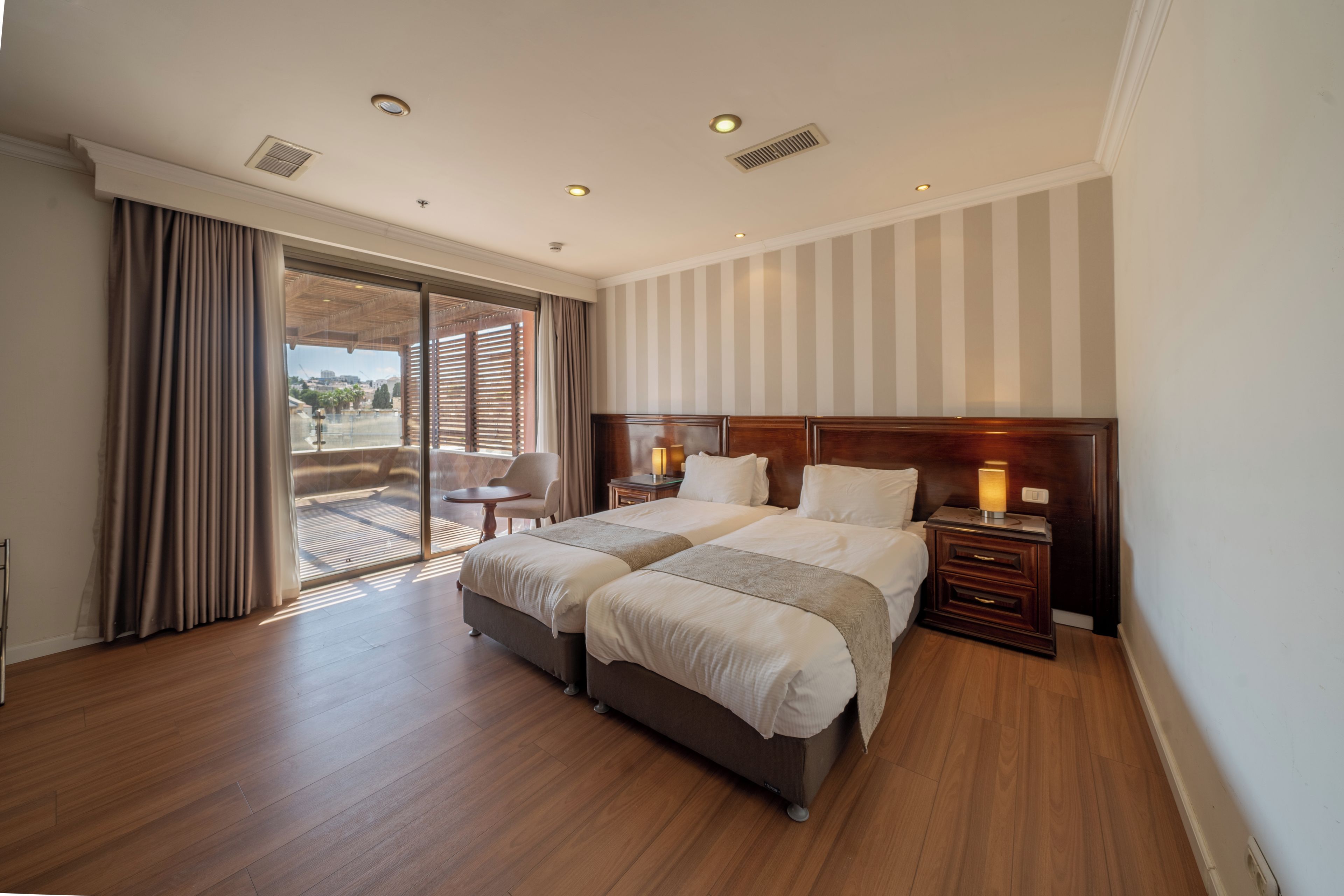 A hotel room with two beds, a wooden floor, a large window with curtains, a view of the outside, and a nightstand with a lamp.