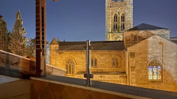 Vista de un edificio de piedra con una torre, posiblemente una iglesia o estructura histórica, visible desde un balcón de madera con barandilla de metal.