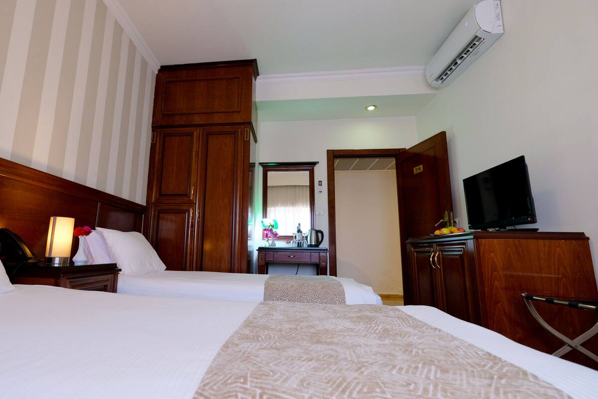 A hotel room with two beds, wooden headboards, a desk with a lamp, a television on a stand, and an air conditioning unit.