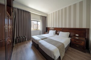 Habitación de hotel con dos camas, cabecero de madera, ventana con cortinas, suelo de madera y un armario.