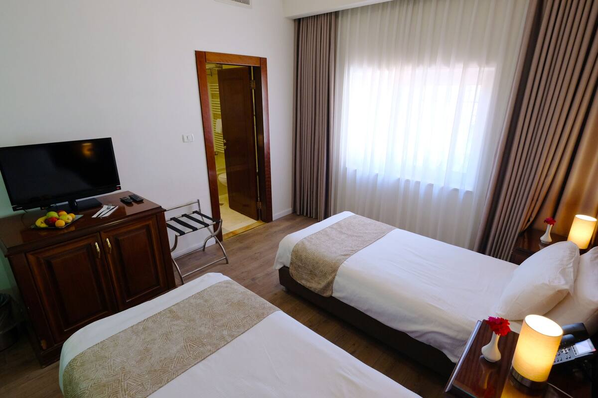 A hotel room with two beds, a TV, a desk with a fruit bowl, and a bathroom door.