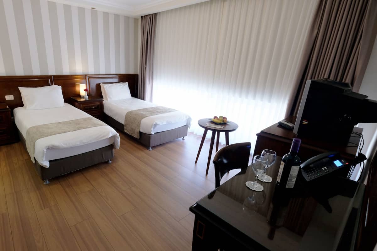 A hotel room with two beds, a desk with a computer and phone, a small table with fruits, and a TV.