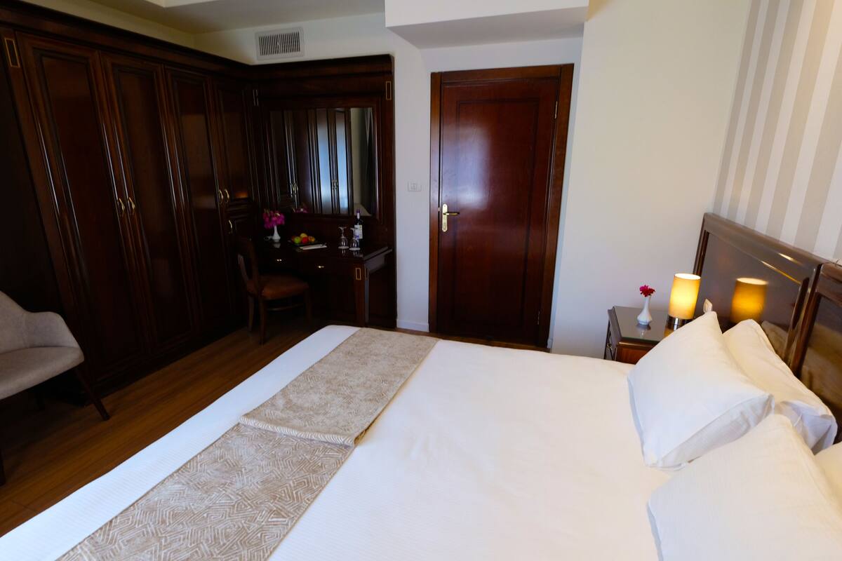 A hotel room with a large bed, a desk with a chair, a wardrobe, and a bedside table with a lamp.