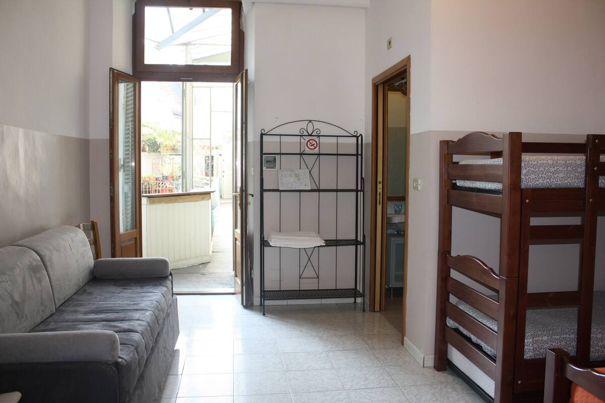 Una habitación con un sofá gris, una reja metálica con un letrero de 'prohibido el paso' y una cama litera de madera.