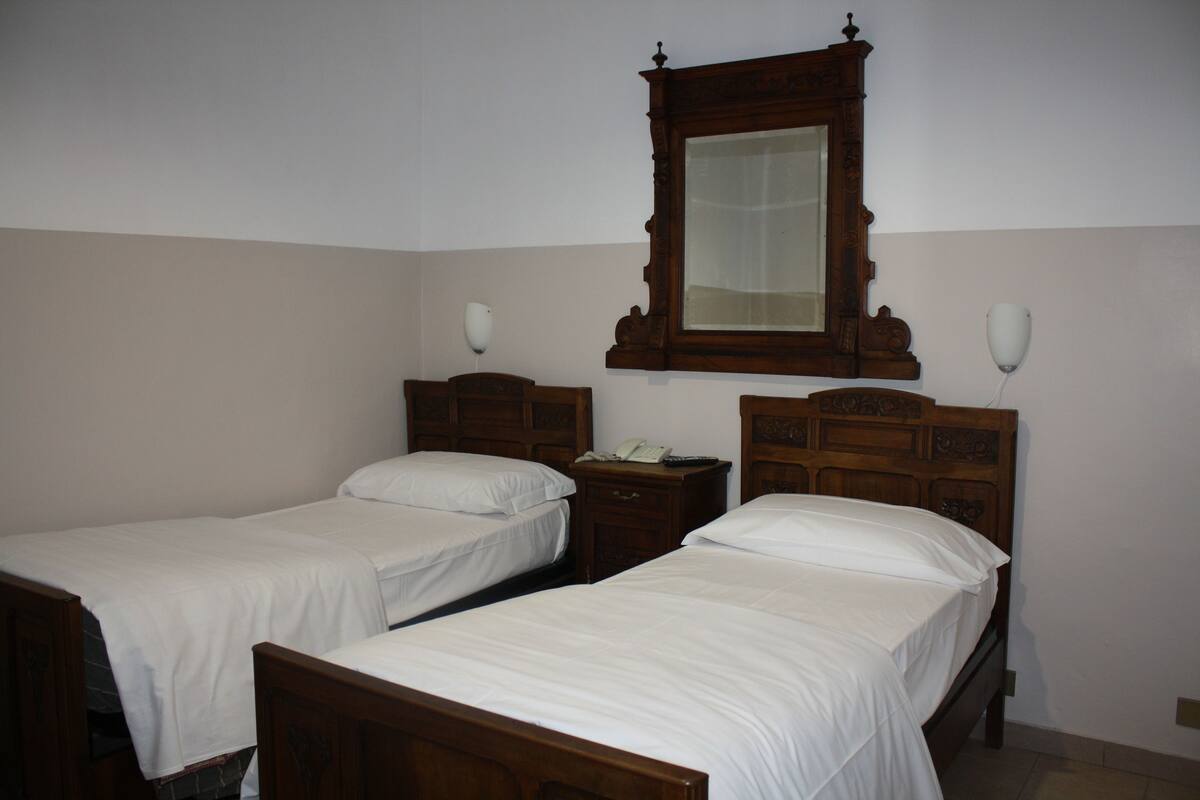 Una habitación con dos camas de madera, cada una con ropa blanca. Hay una mesita de noche de madera con un teléfono y un espejo sobre las camas.