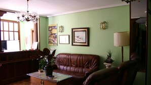 A living room with a brown leather sofa, a glass coffee table, and a wooden cabinet. There is a chandelier, a framed picture on the wall, and a lamp on a side table. - Hosteria Peña Sagra (Cillorigo de Liebana)