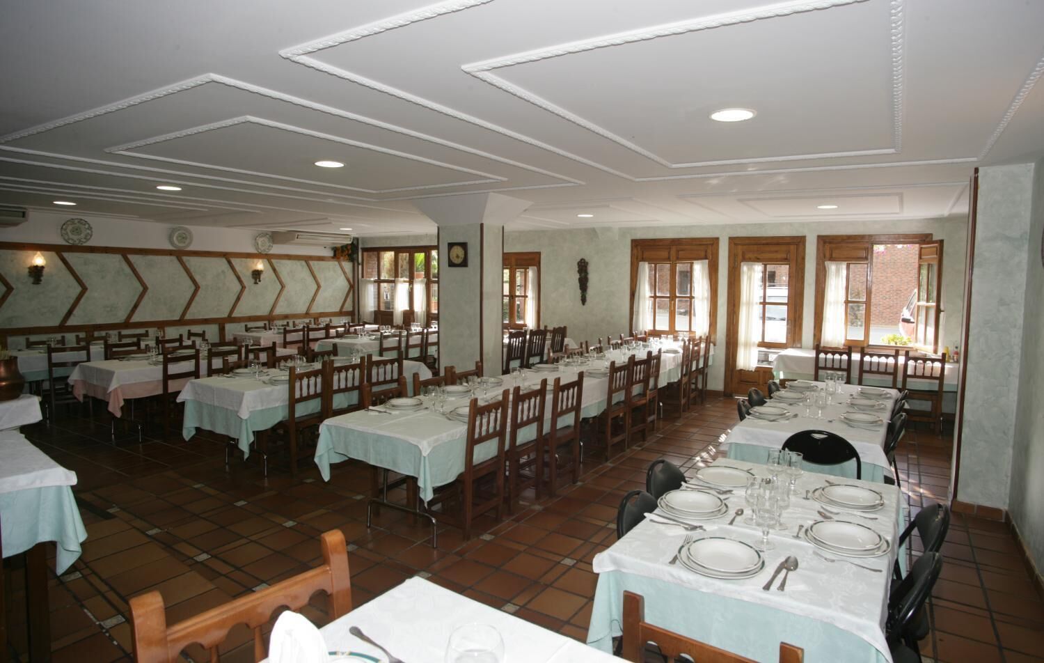 A large dining hall with multiple tables set for meals, featuring a classic design with wooden chairs and white tablecloths.