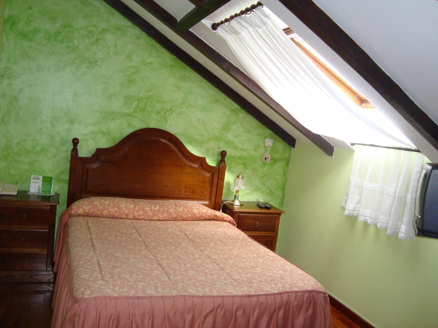 A bedroom with a wooden bed, green walls, a sloped ceiling, and a window with white curtains.