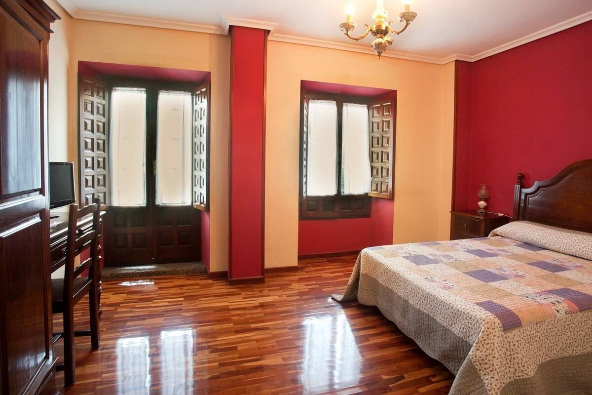 A bedroom with a wooden floor, a bed with a quilt, two windows with curtains, a wooden wardrobe, and a chandelier.