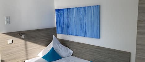 Quarto de hotel moderno com cabeceira de madeira, cama com roupa de cama branca e azul, um quadro abstrato azul na parede e uma pequena mesa de cabeceira com um abajur.