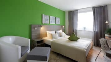 A modern hotel room with a green wall, a large bed with a green pillow, a white armchair, a small table, and a nightstand.