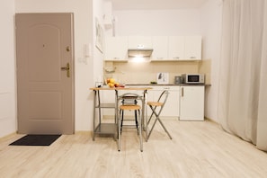 Μια μικρή, λειτουργική κουζίνα με λευκά ντουλάπια, φούρνο μικροκυμάτων, νεροχύτη και ένα μικρό χώρο τραπεζαρίας με σκαμπό.
