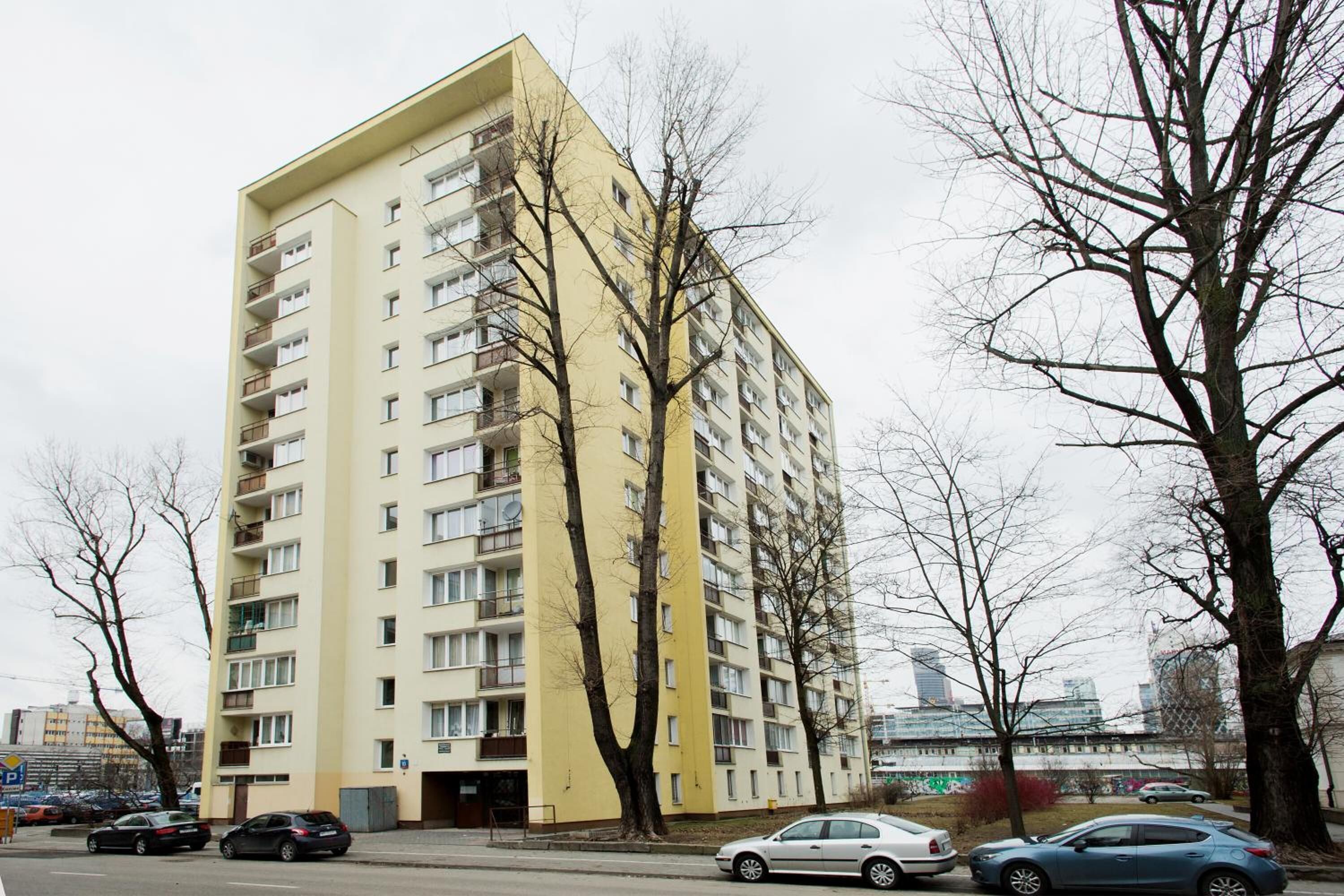 A multi-story apartment building with balconies, parked cars, and leafless trees.
