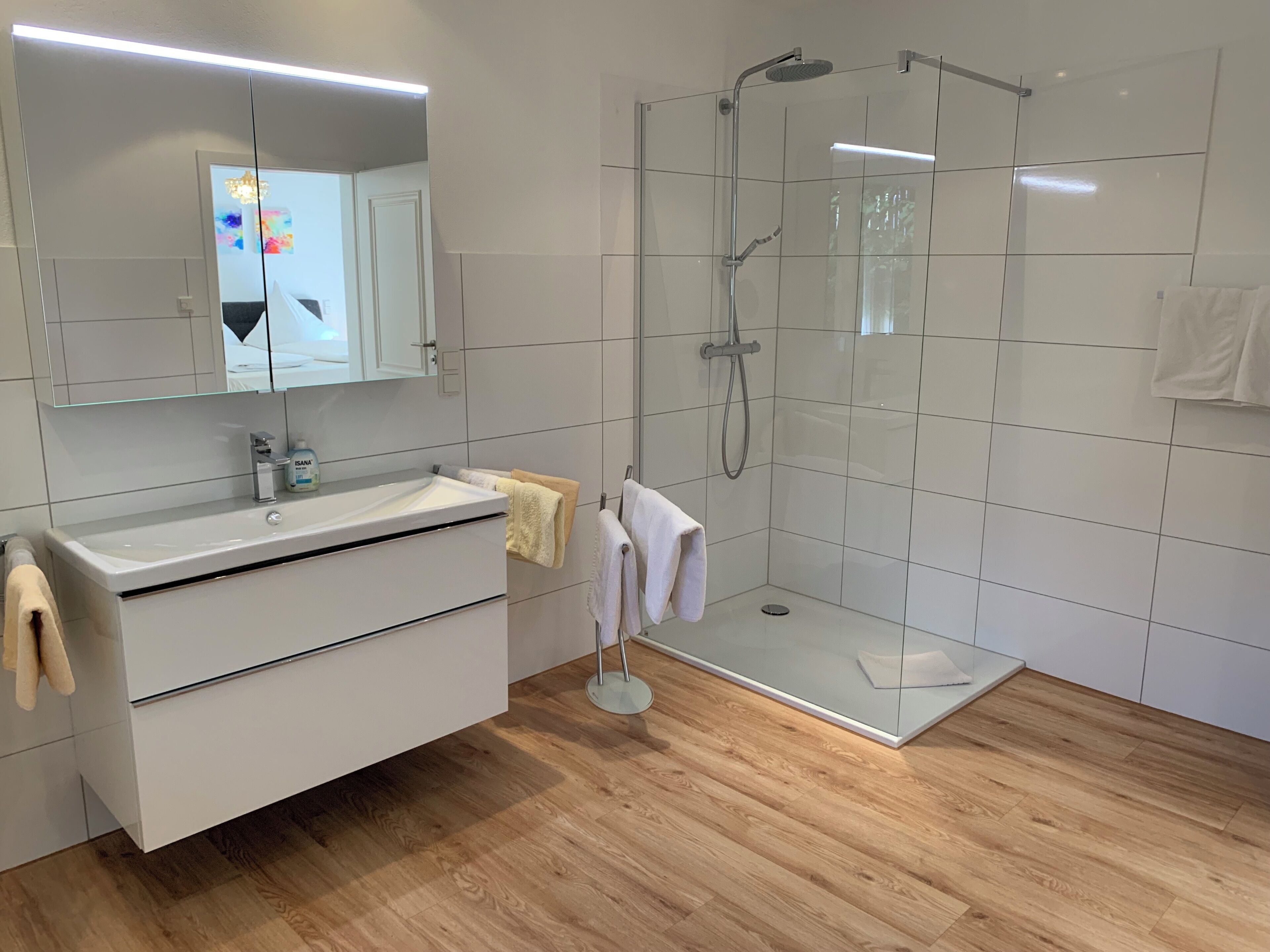 Een moderne badkamer met een douchecabine van glas, een witte wastafel met spiegel, en een houten vloer.