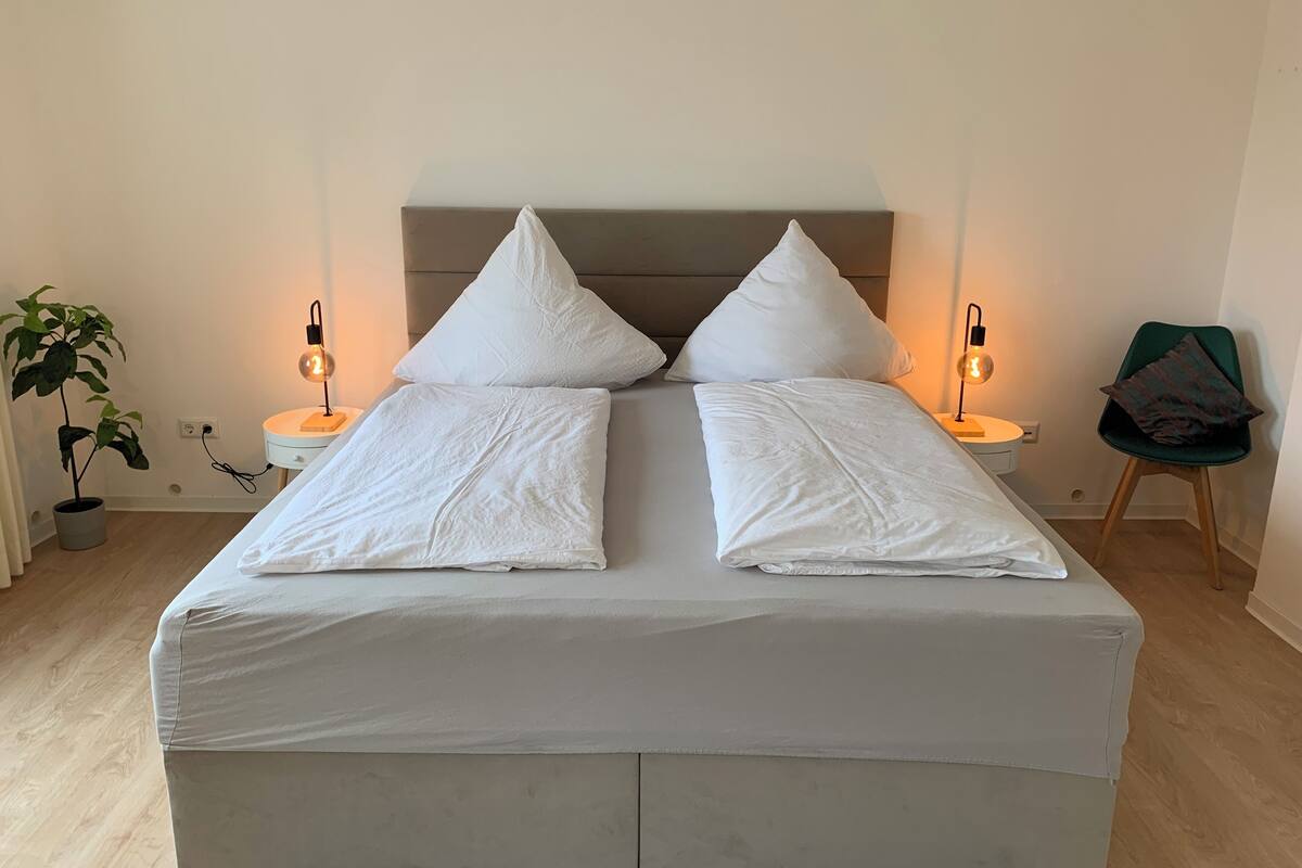 Un lit avec deux oreillers, deux tables de chevet avec des lampes, une plante en pot et une chaise.