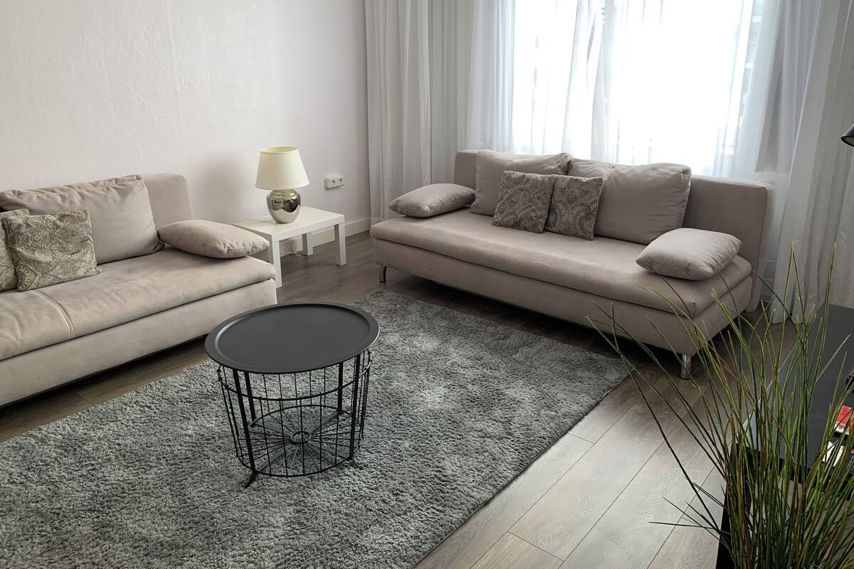 Un salon moderne avec deux canapés, une petite table d’appoint, une table basse ronde et une grande fenêtre agrémentée de rideaux légers.