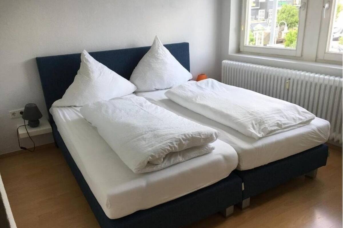Un lit avec une literie blanche et une tête de lit sombre, dans une pièce dotée d’une fenêtre et d’un radiateur.