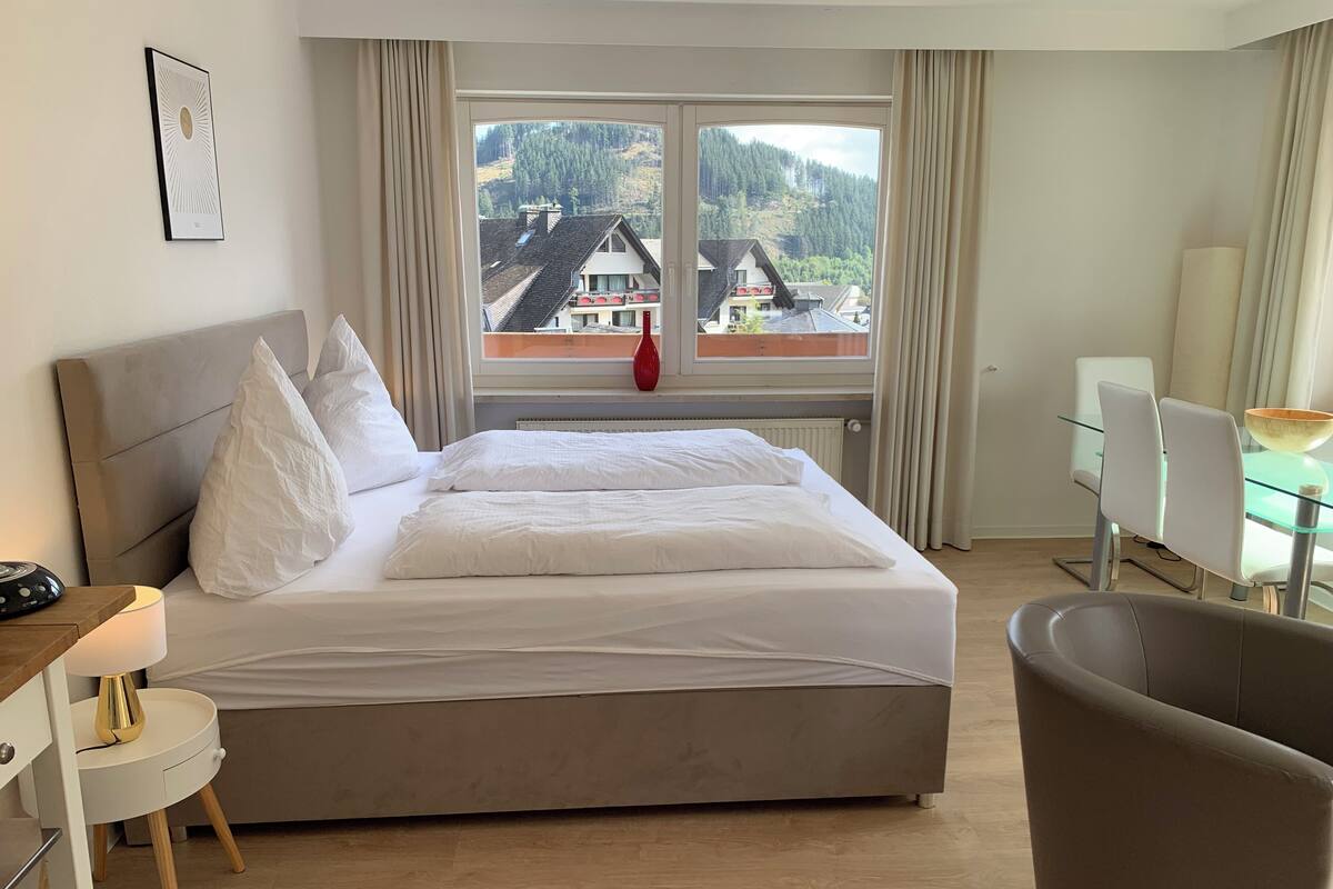 Une chambre d’hôtel avec un lit, un bureau et une chaise. Il y a une fenêtre donnant sur des immeubles et des arbres.