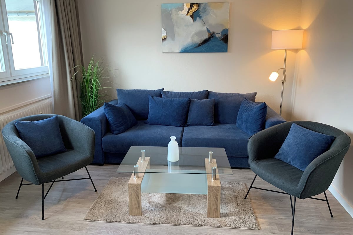 Un salon moderne avec un canapé bleu foncé, deux fauteuils, une table basse en verre et un tableau accroché au mur.
