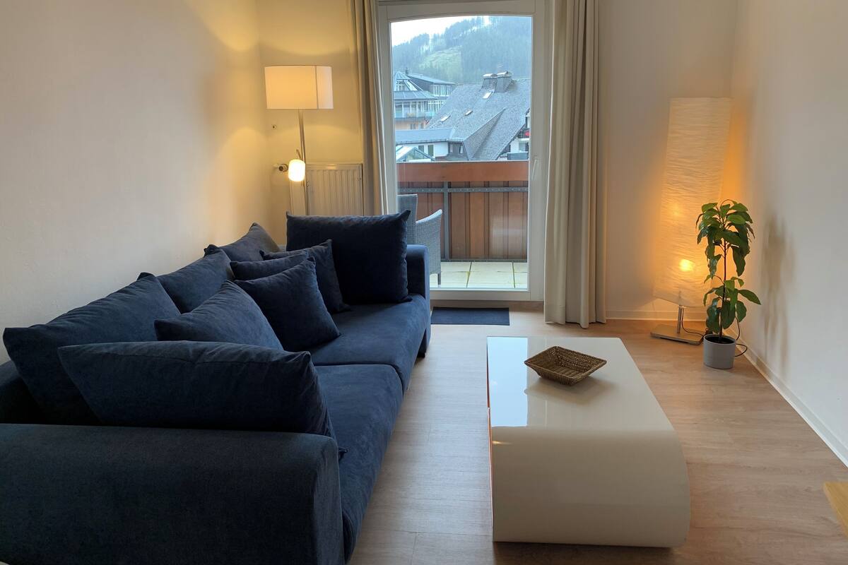Un salon moderne avec un canapé bleu foncé, un pouf blanc et une plante en pot. Une grande fenêtre offre une vue sur un bâtiment doté d’un toit en pente.
