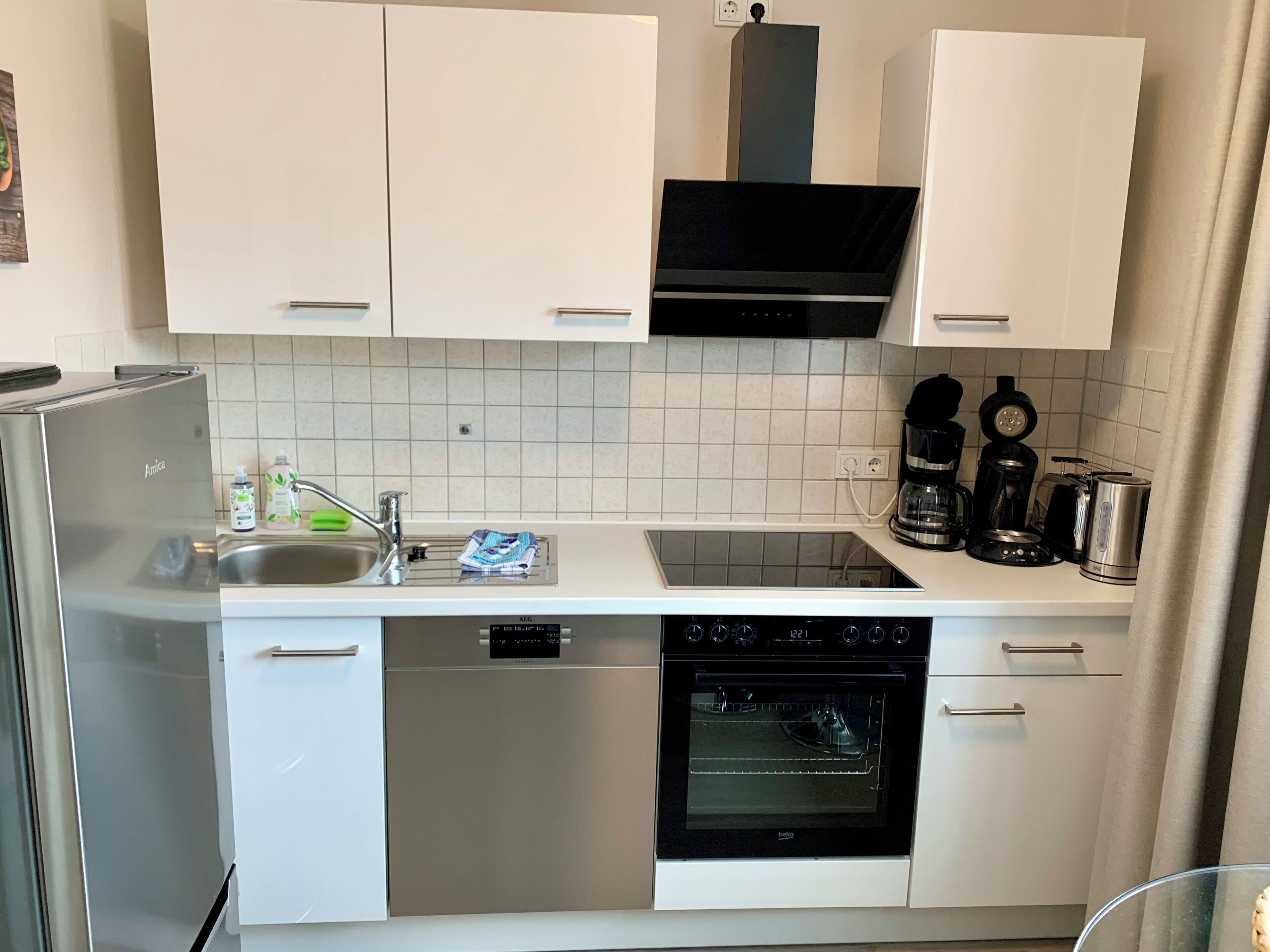 Una cuina compacta amb armaris blancs, un frigorífic d'acer inoxidable, un rentaplats, un forn i una cafetera.