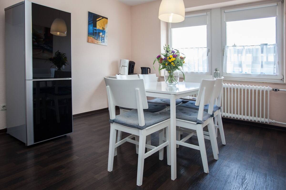 Une cuisine moderne avec un coin repas, des meubles blancs, une machine à café et une fenêtre donnant sur un beau paysage.