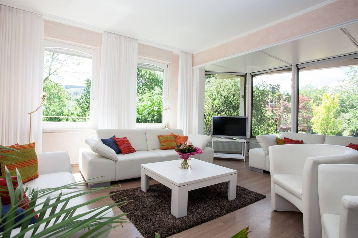 Un salon moderne avec un ensemble de canapés blancs, une table basse et un téléviseur. De grandes fenêtres offrent une vue sur la verdure environnante.