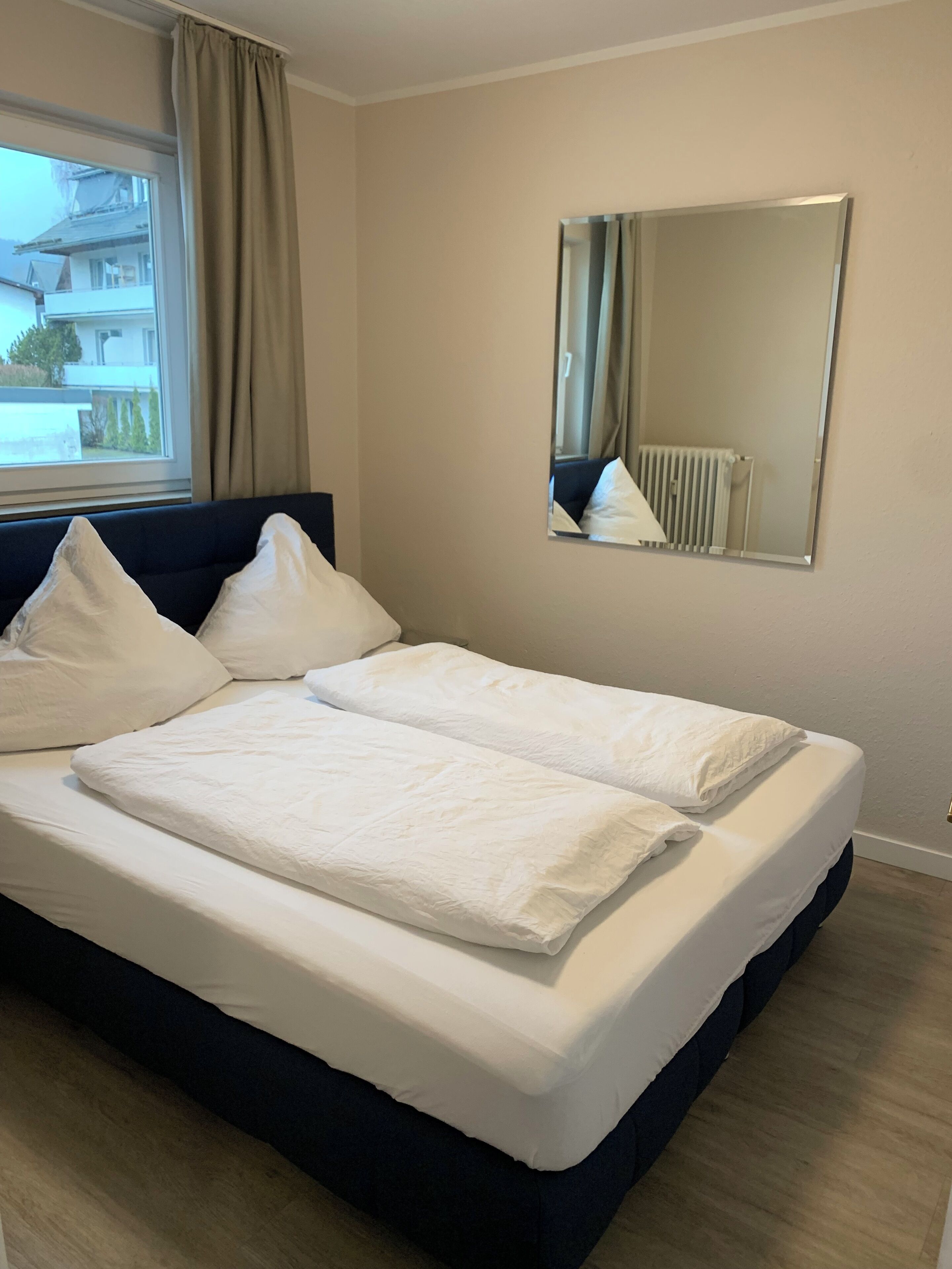 Ein ordentlich bezogenes Bett mit weißen Bettwäsche in einem Zimmer mit einem großen Fenster, einem Spiegel an der Wand und Blick auf Gebäude im Freien.