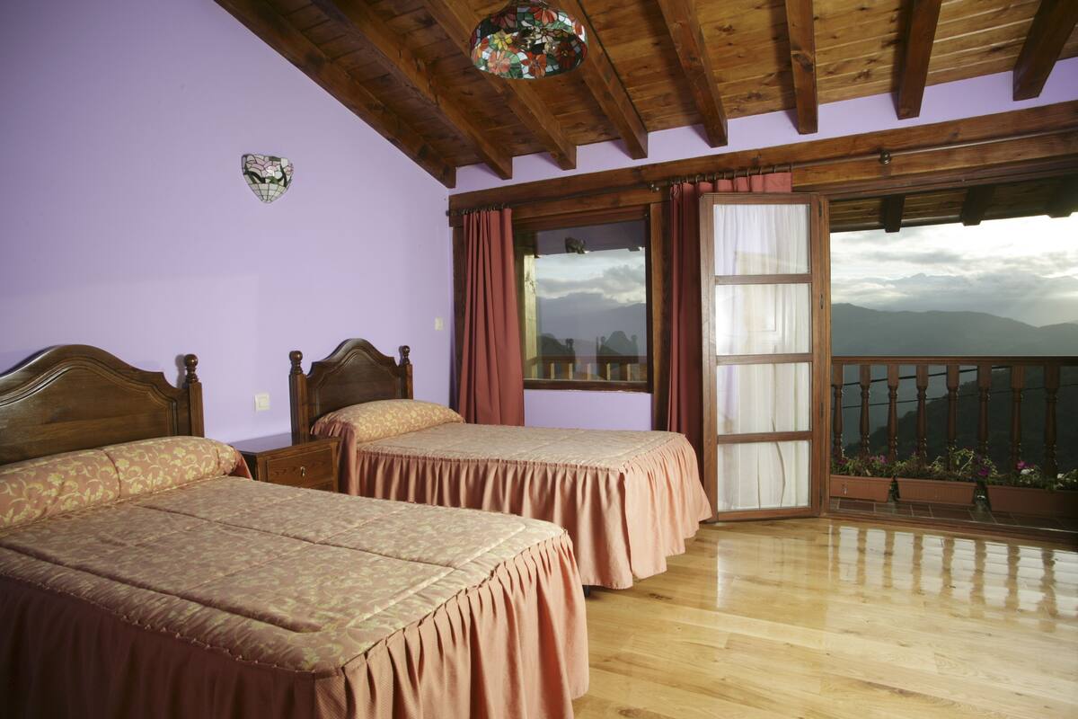 A room with two wooden beds, a wooden ceiling, a large window with curtains, and a balcony with a view of mountains.